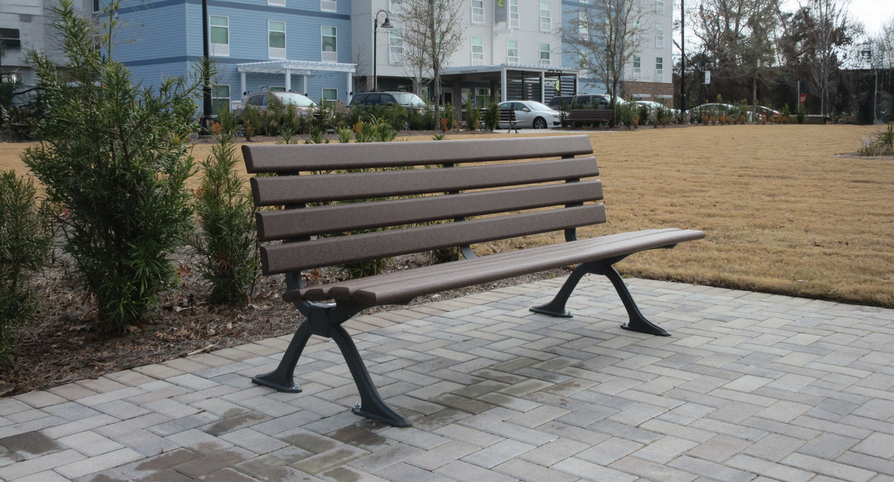 A brown wooden park bench with metal legs sits on a paved area near a grassy field, with shrubs and buildings in the background.