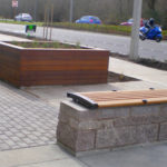 A sidewalk with a wooden bench, a large rectangular wooden planter, and a street with passing cars and a motorcyclist in the background.