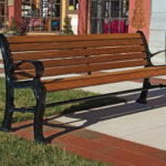A wooden and metal bench is placed on a sidewalk next to a patch of grass, in front of storefronts with large windows.