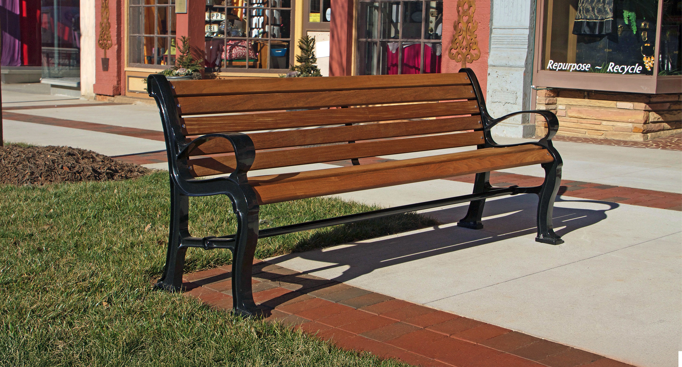maglin_mbe-0300-backed-benches-1 A wooden and metal bench is placed on a sidewalk next to a patch of grass, in front of storefronts with large windows.