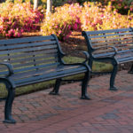 Two empty dark green metal benches on a brick walkway in a park, with blooming pink flowers and green shrubs in the background.