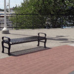Metal benches on a paved promenade by a waterfront, with boats, a tree, and railings in the background.
