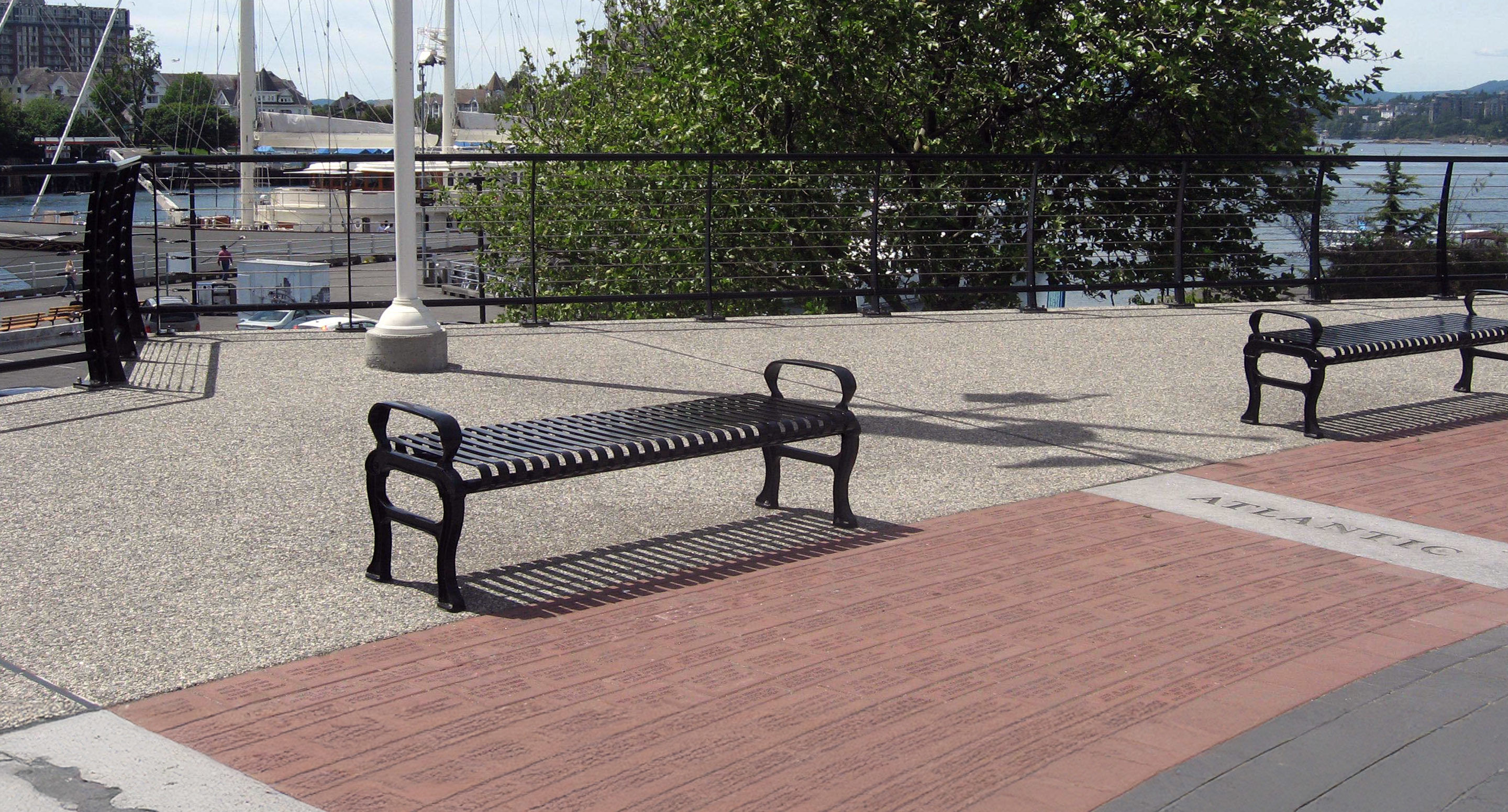 Metal benches on a paved promenade by a waterfront, with boats, a tree, and railings in the background.