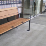 Deux bancs en bois avec des cadres en métal noir sont placés à l'extérieur d'un bâtiment avec des portes et des fenêtres en verre, sur un trottoir en béton.