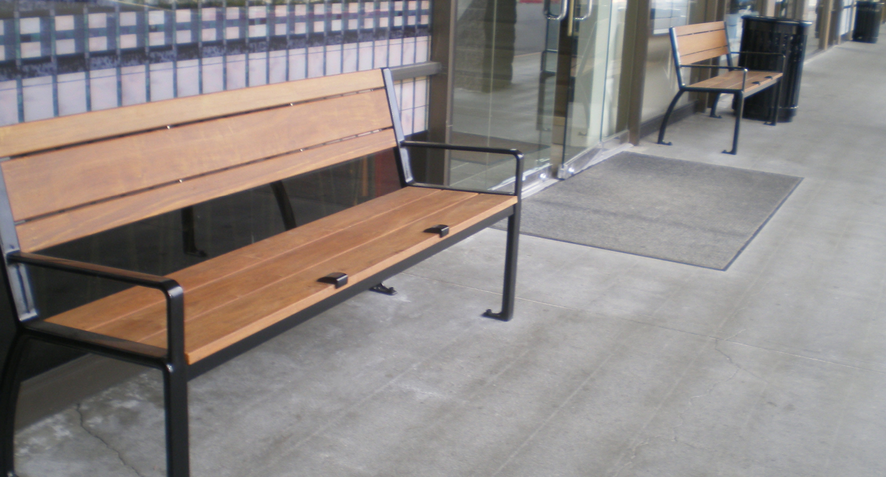 maglin_mbe-0300-bench-skate-deterrents-1 Deux bancs en bois avec des cadres en métal noir sont placés à l'extérieur d'un bâtiment avec des portes et des fenêtres en verre, sur un trottoir en béton.