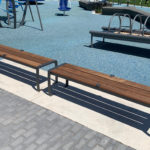 Deux bancs en bois avec des armatures métalliques sont placés sur une dalle de béton à côté d'une aire de jeux caoutchoutée avec des équipements de jeux à l'arrière-plan.