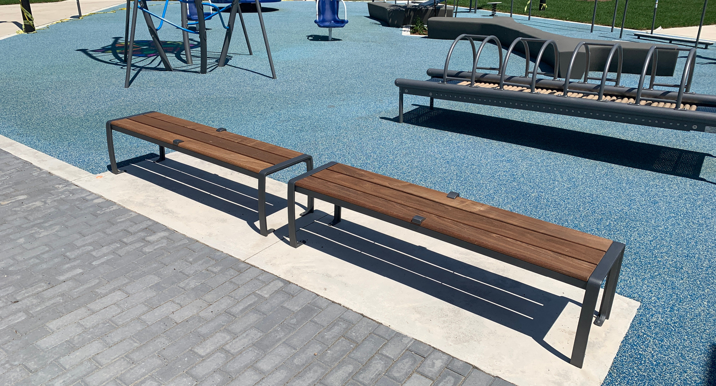 maglin_mbe-0300-bench-skate-deterrents-2 Deux bancs en bois avec des armatures métalliques sont placés sur une dalle de béton à côté d'une aire de jeux caoutchoutée avec des équipements de jeux à l'arrière-plan.
