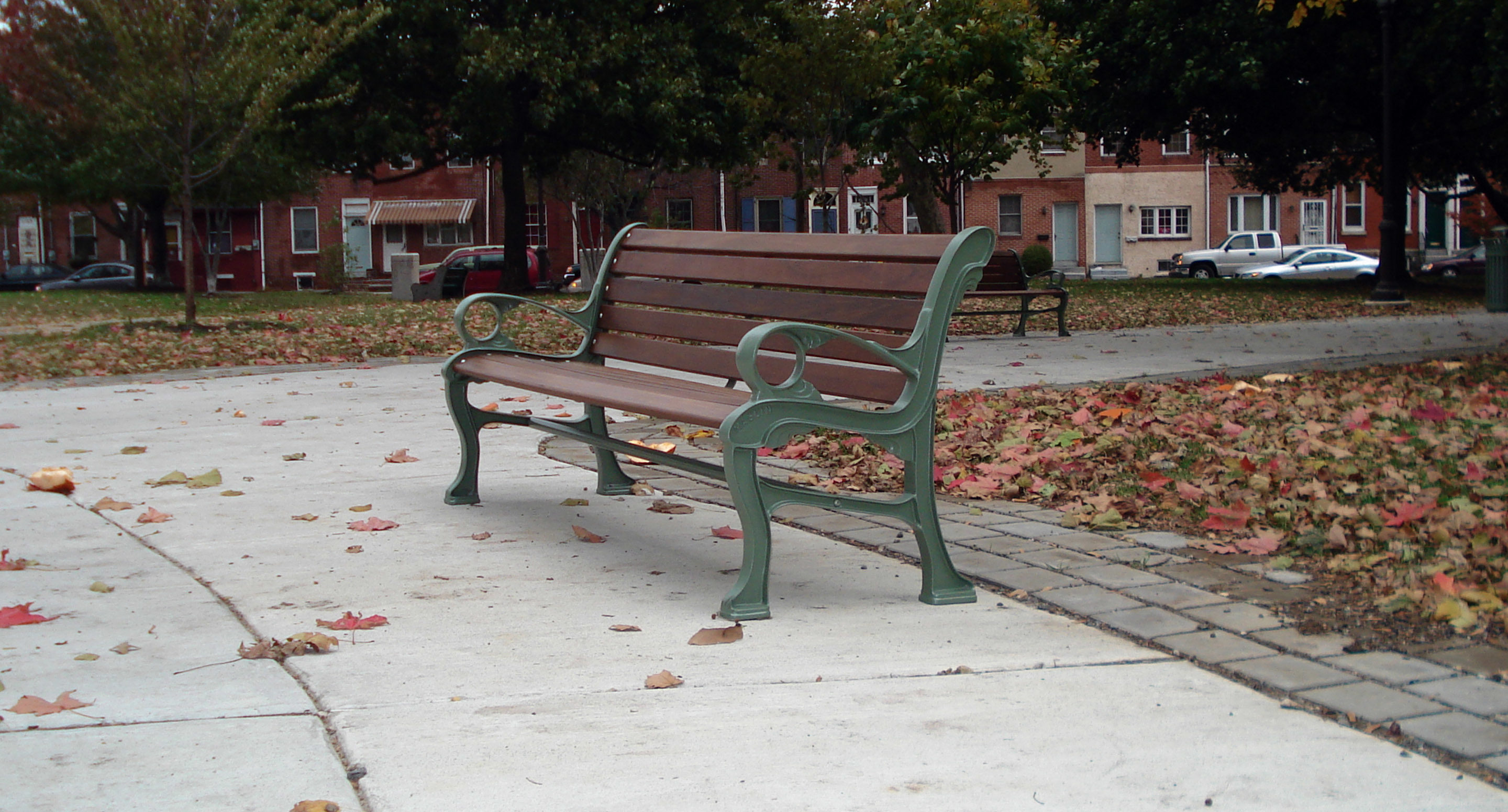 A wooden park bench with metal armrests sits on a paved path surrounded by fallen leaves and trees in an urban park.