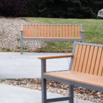 Two empty wooden benches with metal frames are placed on a paved path in a landscaped outdoor area with grass, mulch, and greenery.