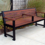 A wooden and metal bench with a center armrest is placed on a sidewalk next to a building and landscaping.