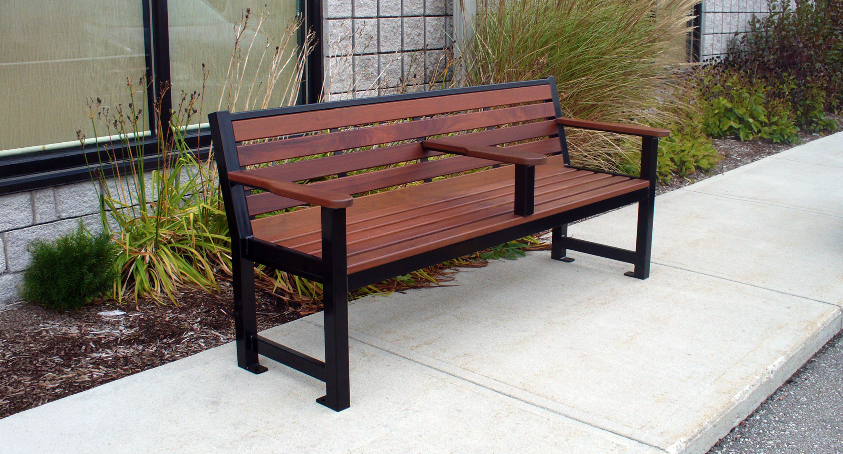 maglin_mbe-0400-backed-benches-1 A wooden and metal bench with a center armrest is placed on a sidewalk next to a building and landscaping.