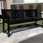 Black metal bench on a stone-paved walkway with artificial grass and trimmed shrubs in the background, next to a building.