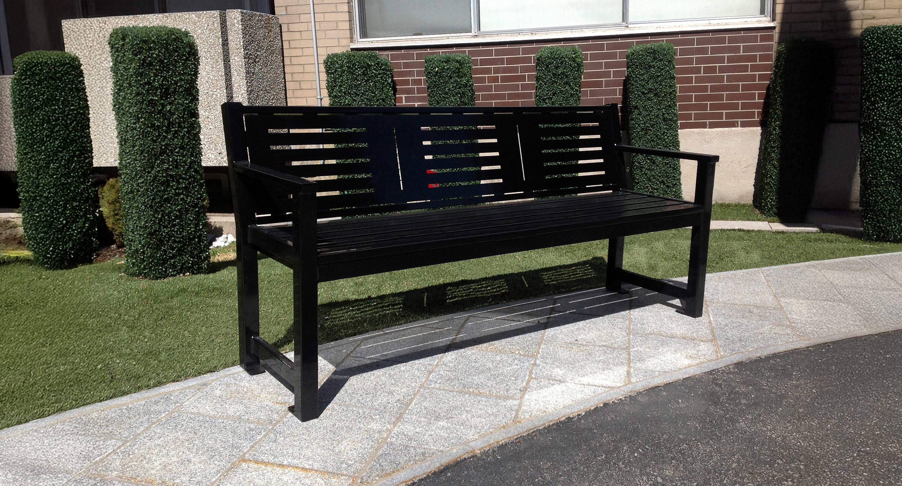 Black metal bench on a stone-paved walkway with artificial grass and trimmed shrubs in the background, next to a building.