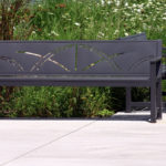 Two metal benches with decorative cutouts are placed on a paved area in front of tall green grass and white wildflowers.