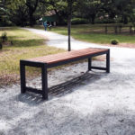 A wooden bench with metal legs is placed on a gravel path in a park, surrounded by grass, trees, and a winding walkway.
