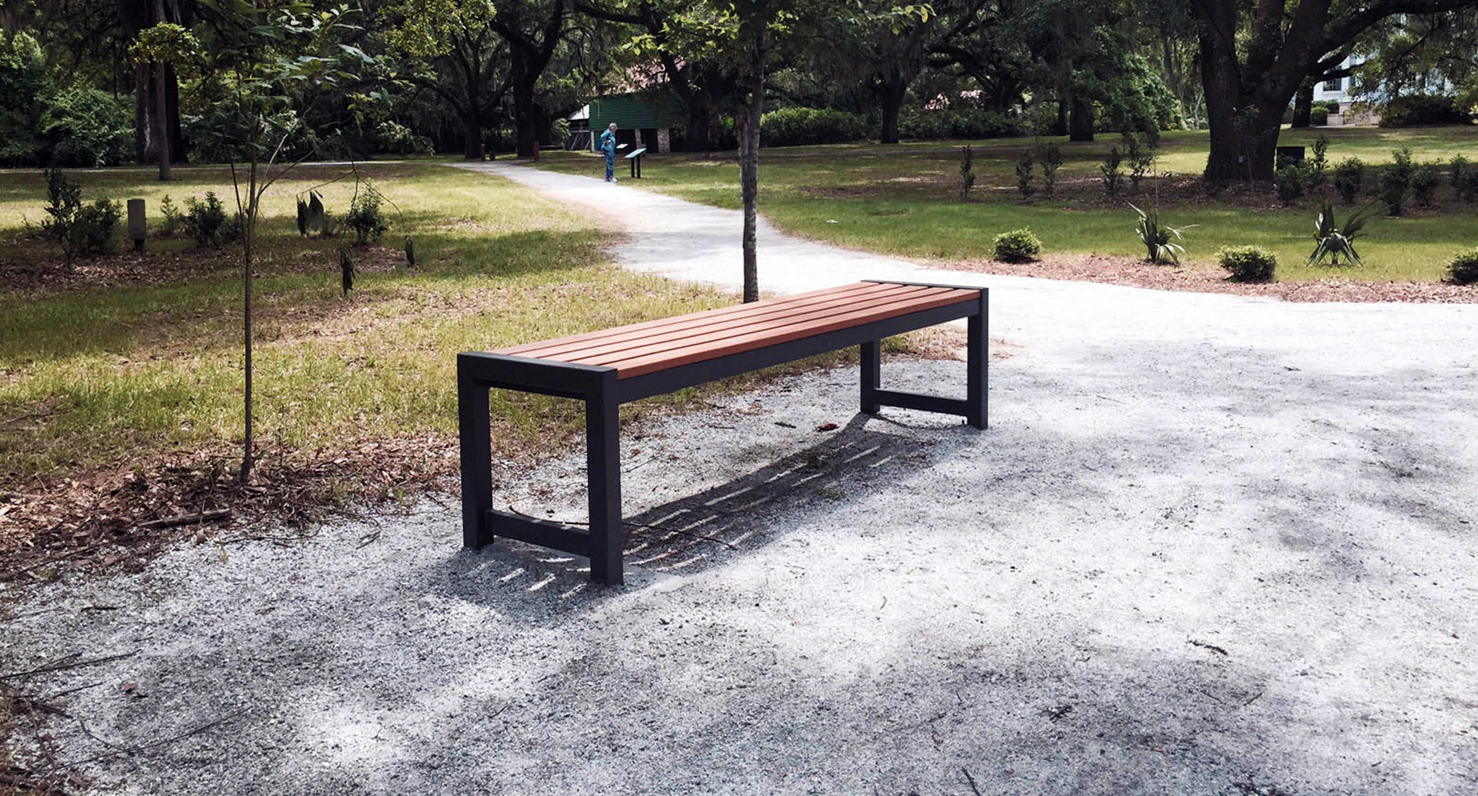 A wooden bench with metal legs is placed on a gravel path in a park, surrounded by grass, trees, and a winding walkway.