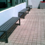 Three metal benches are lined up on a tiled outdoor patio next to a building with large windows; a planter with red flowers is visible in the background.