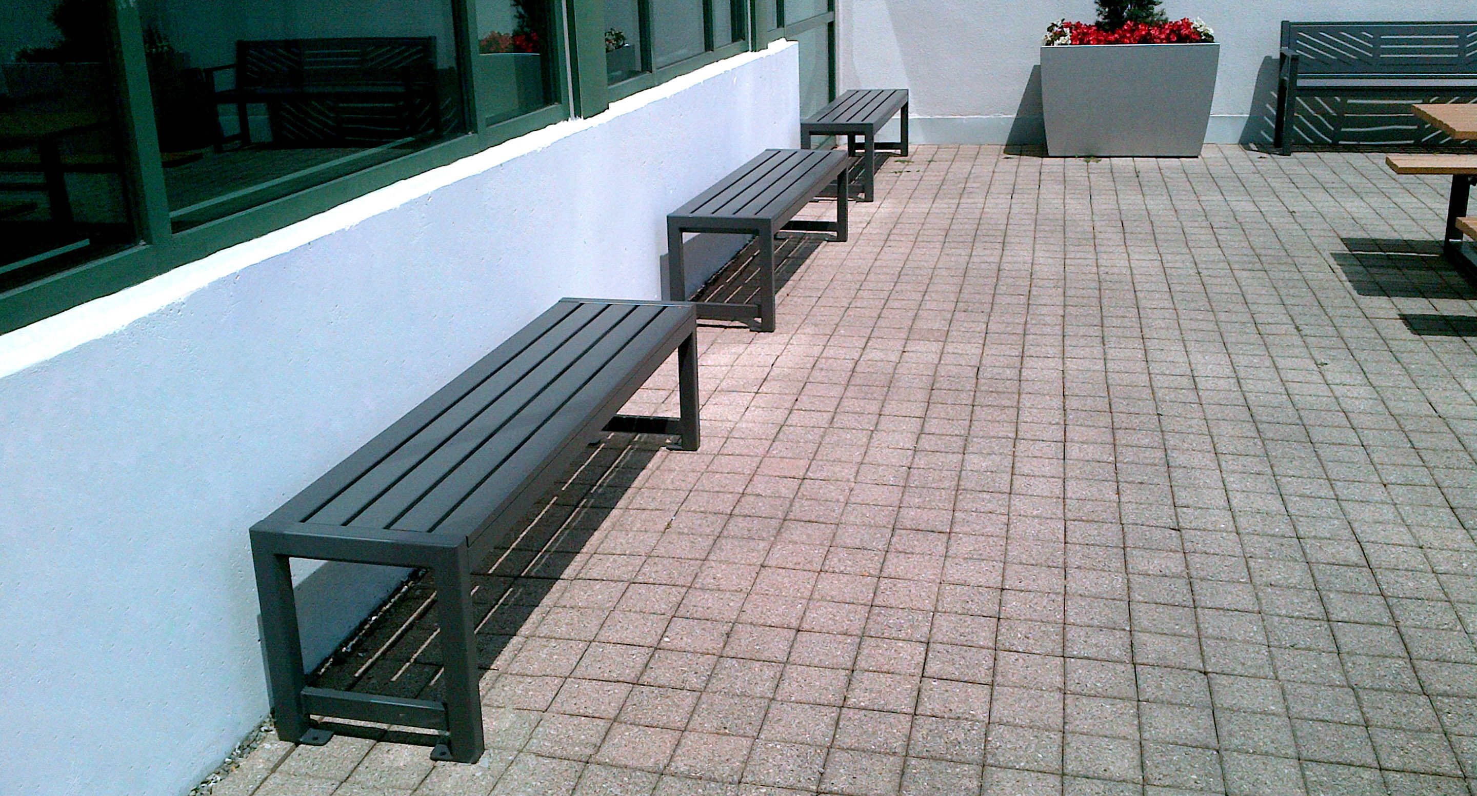 Three metal benches are lined up on a tiled outdoor patio next to a building with large windows; a planter with red flowers is visible in the background.