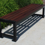 A modern outdoor bench with a dark wooden seat and black metal frame sits on a concrete sidewalk next to green plants.