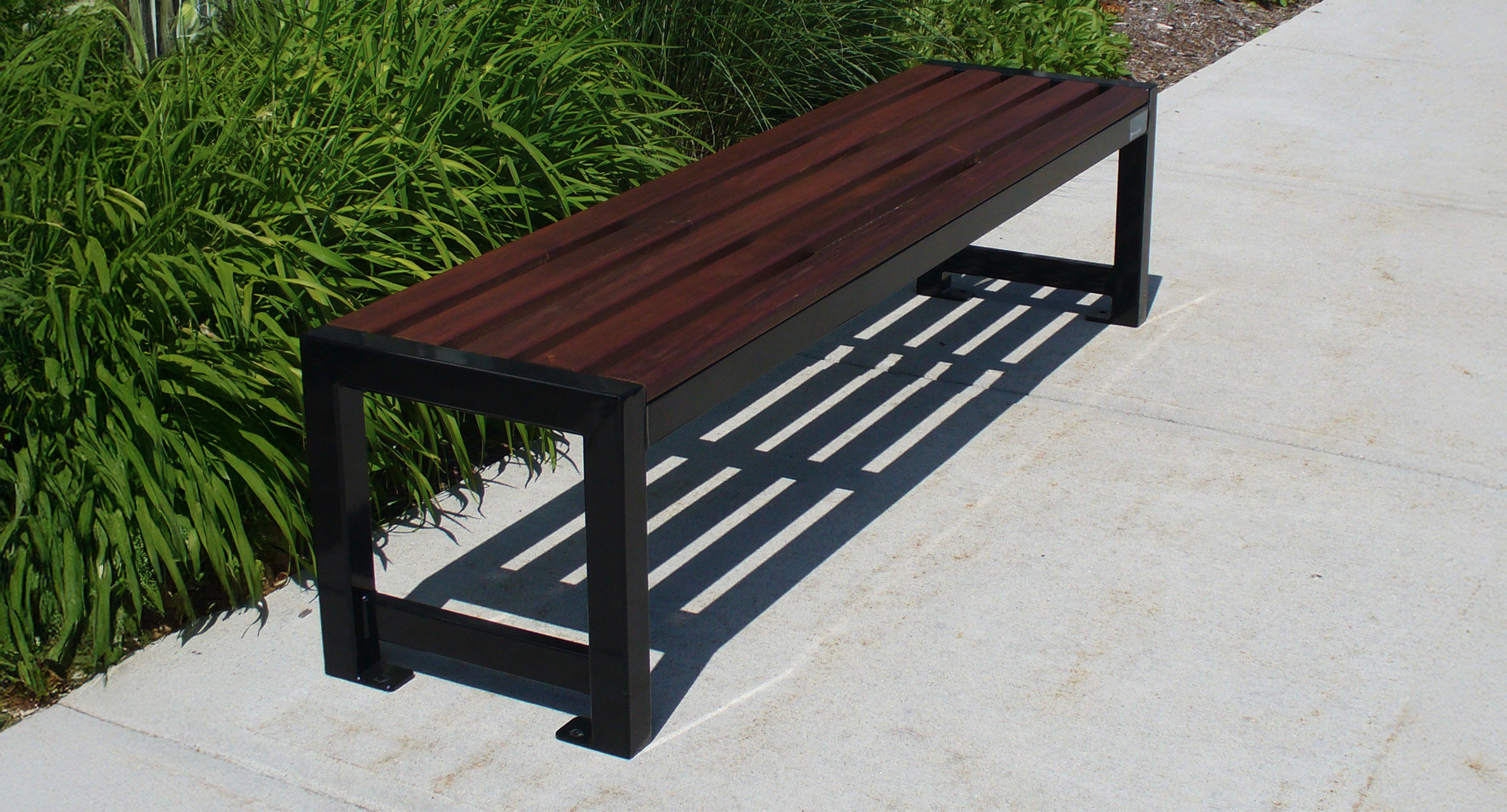 A modern outdoor bench with a dark wooden seat and black metal frame sits on a concrete sidewalk next to green plants.