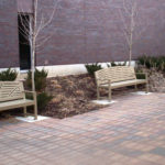 Three beige metal benches are placed along a brick sidewalk next to a landscaped area with small shrubs and leafless trees in front of a brick building.