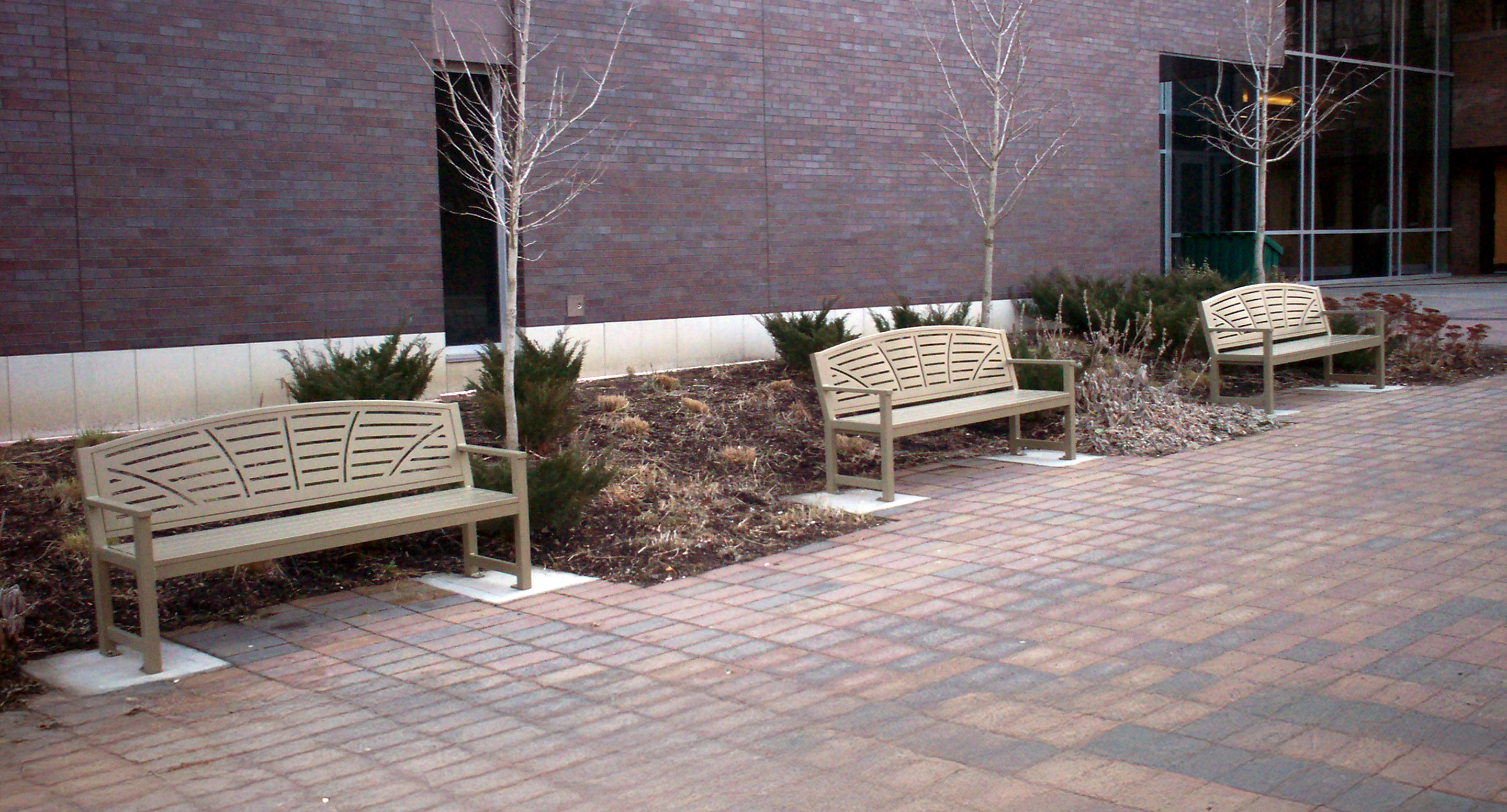 maglin_mbe-0450-backed-benches-1 Three beige metal benches are placed along a brick sidewalk next to a landscaped area with small shrubs and leafless trees in front of a brick building.