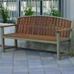 A wooden bench with armrests is placed on a tiled pavement near a concrete planter with green plants, next to a metal fence.