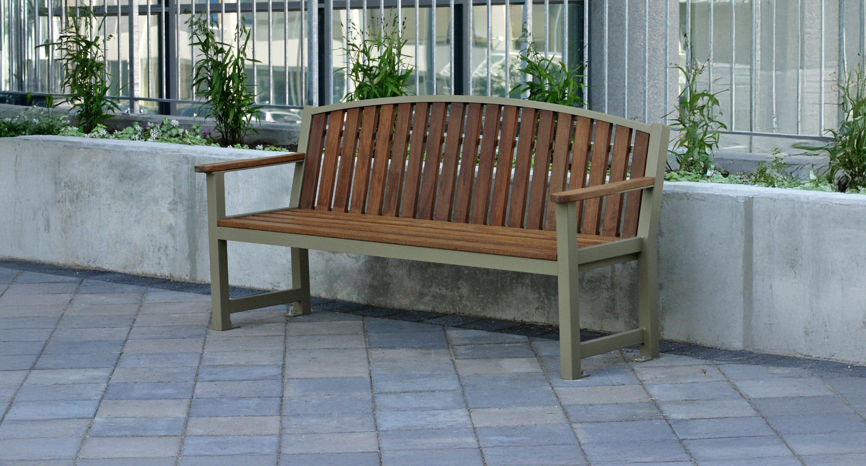 maglin_mbe-0450-backed-benches-2 A wooden bench with armrests is placed on a tiled pavement near a concrete planter with green plants, next to a metal fence.