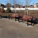 A paved park path with wooden benches, metal tables, a trash can, small trees, and houses in the background under a clear sky.