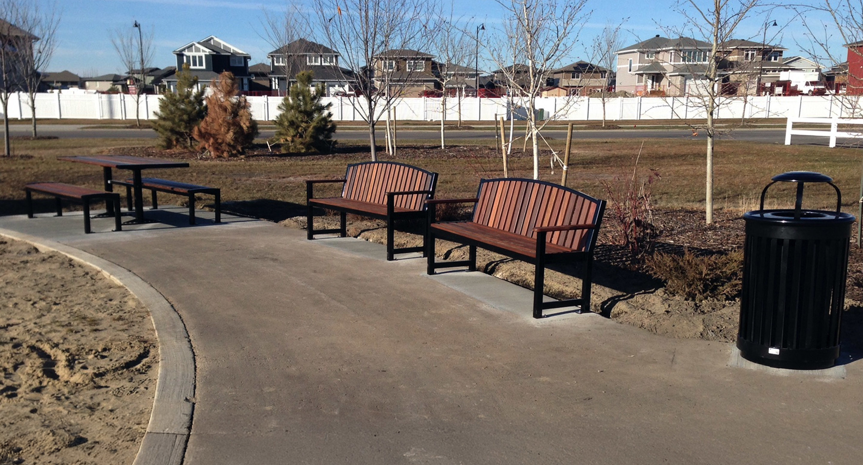 maglin_mbe-0450-backed-benches-3 A paved park path with wooden benches, metal tables, a trash can, small trees, and houses in the background under a clear sky.