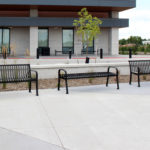 Three black metal benches are arranged on a concrete patio outside a modern building with large windows and landscaping features.