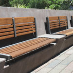 Three wooden benches with metal frames are mounted on a concrete wall along a paved walkway, with greenery visible in the background.