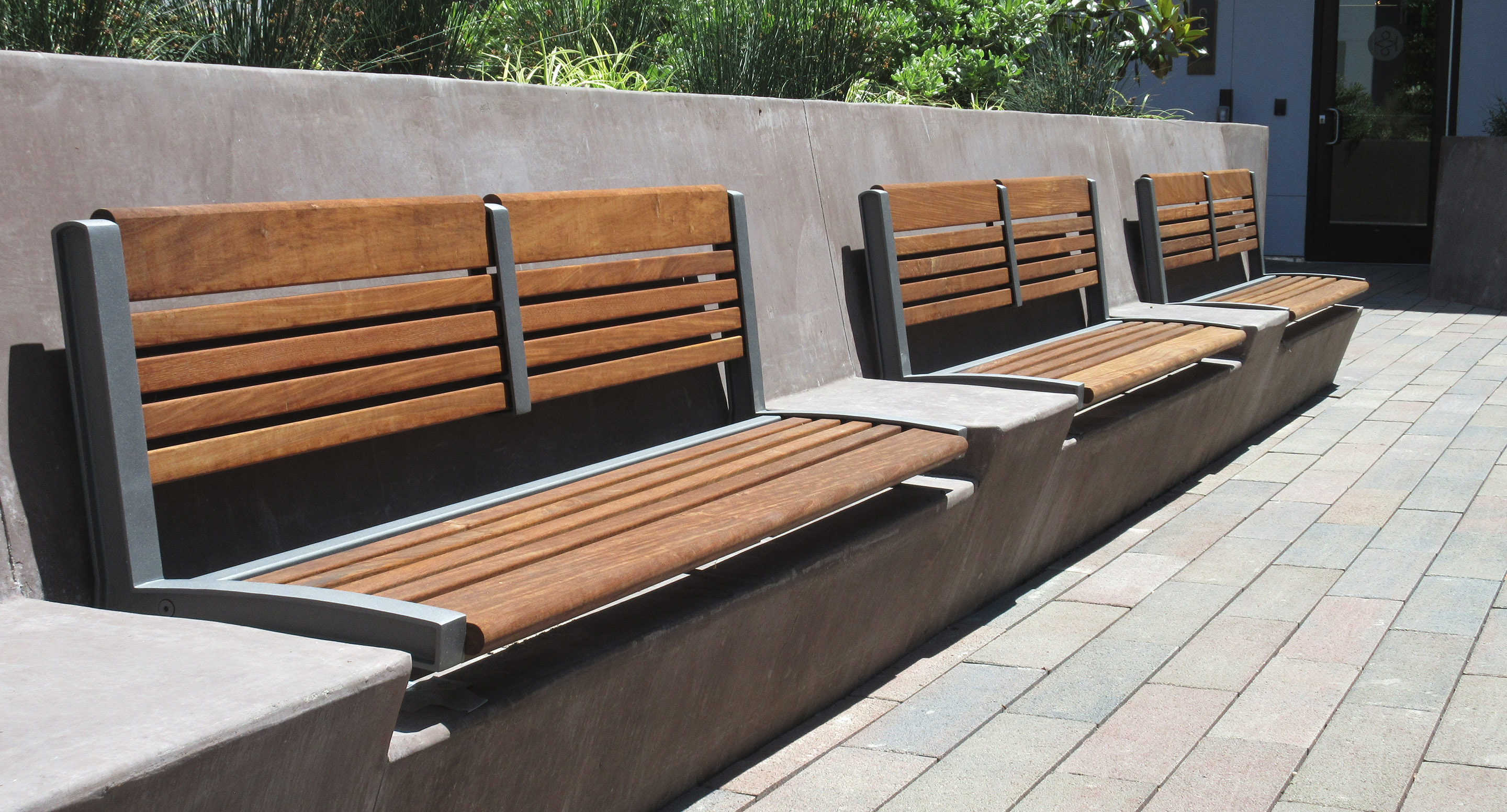 Three wooden benches with metal frames are mounted on a concrete wall along a paved walkway, with greenery visible in the background.