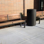 Two wooden benches and a trash can are positioned on a concrete sidewalk next to a brick building.