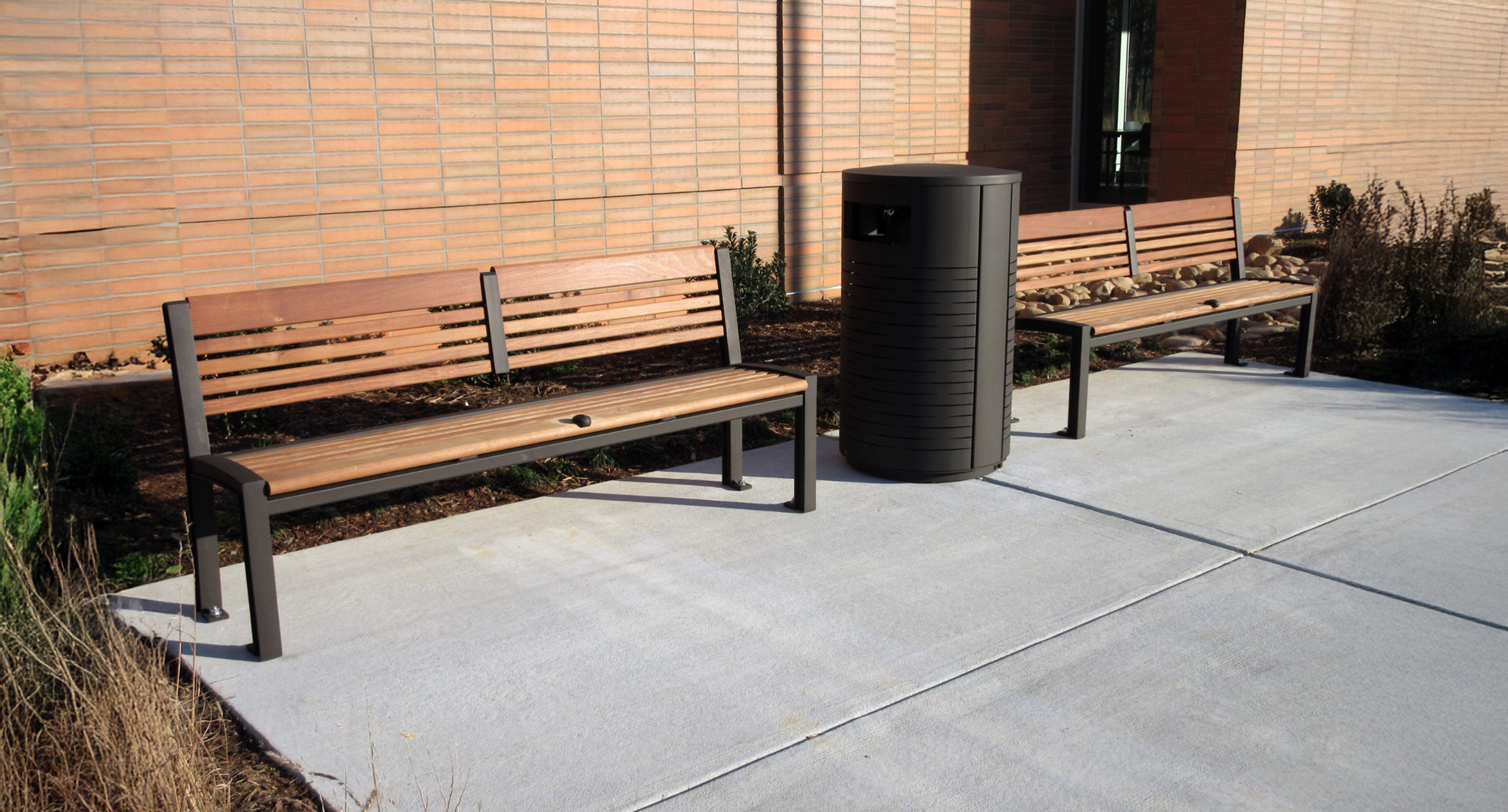 Two wooden benches and a trash can are positioned on a concrete sidewalk next to a brick building.