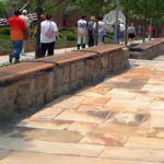Stone pathway with long wooden benches along a stone wall, several people walking in groups toward a modern building in the background.