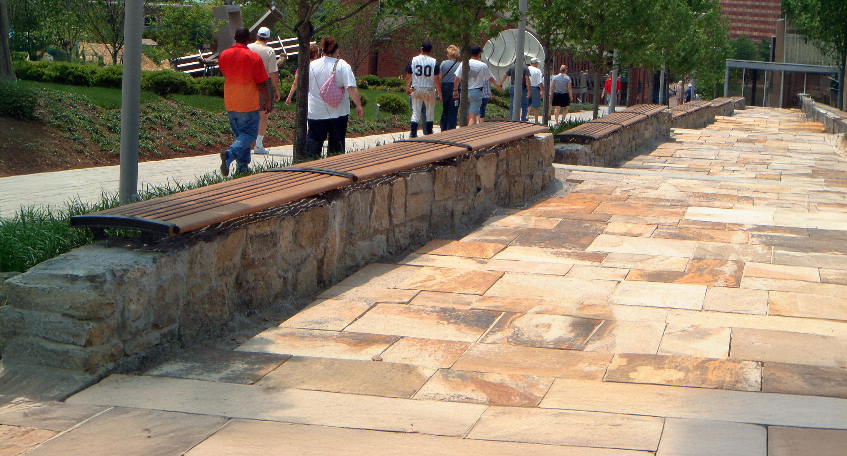 maglin_mbe-0720-backless-benches-1 Stone pathway with long wooden benches along a stone wall, several people walking in groups toward a modern building in the background.