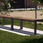 A long wooden bench with a metal frame stands on a paved area surrounded by grass in a park, with trees and people visible in the background.