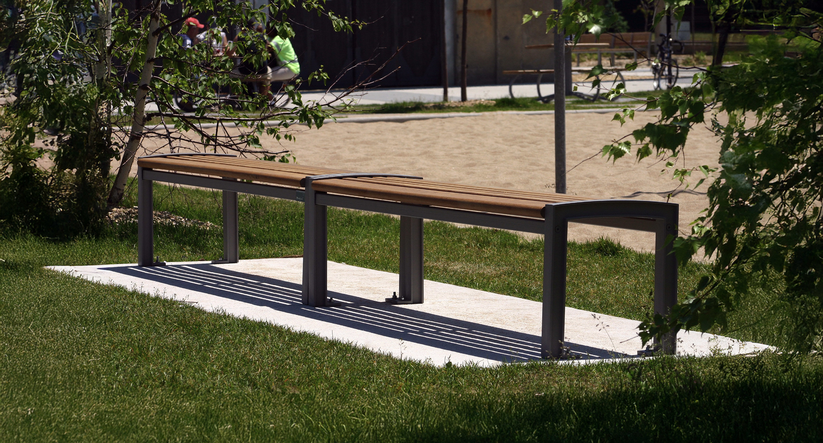 maglin_mbe-0720-backless-benches-2 A long wooden bench with a metal frame stands on a paved area surrounded by grass in a park, with trees and people visible in the background.