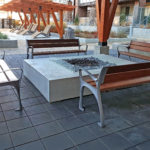 Four wooden benches surround a square concrete fire pit on a tiled patio, with a pergola and landscaped plants in the background.