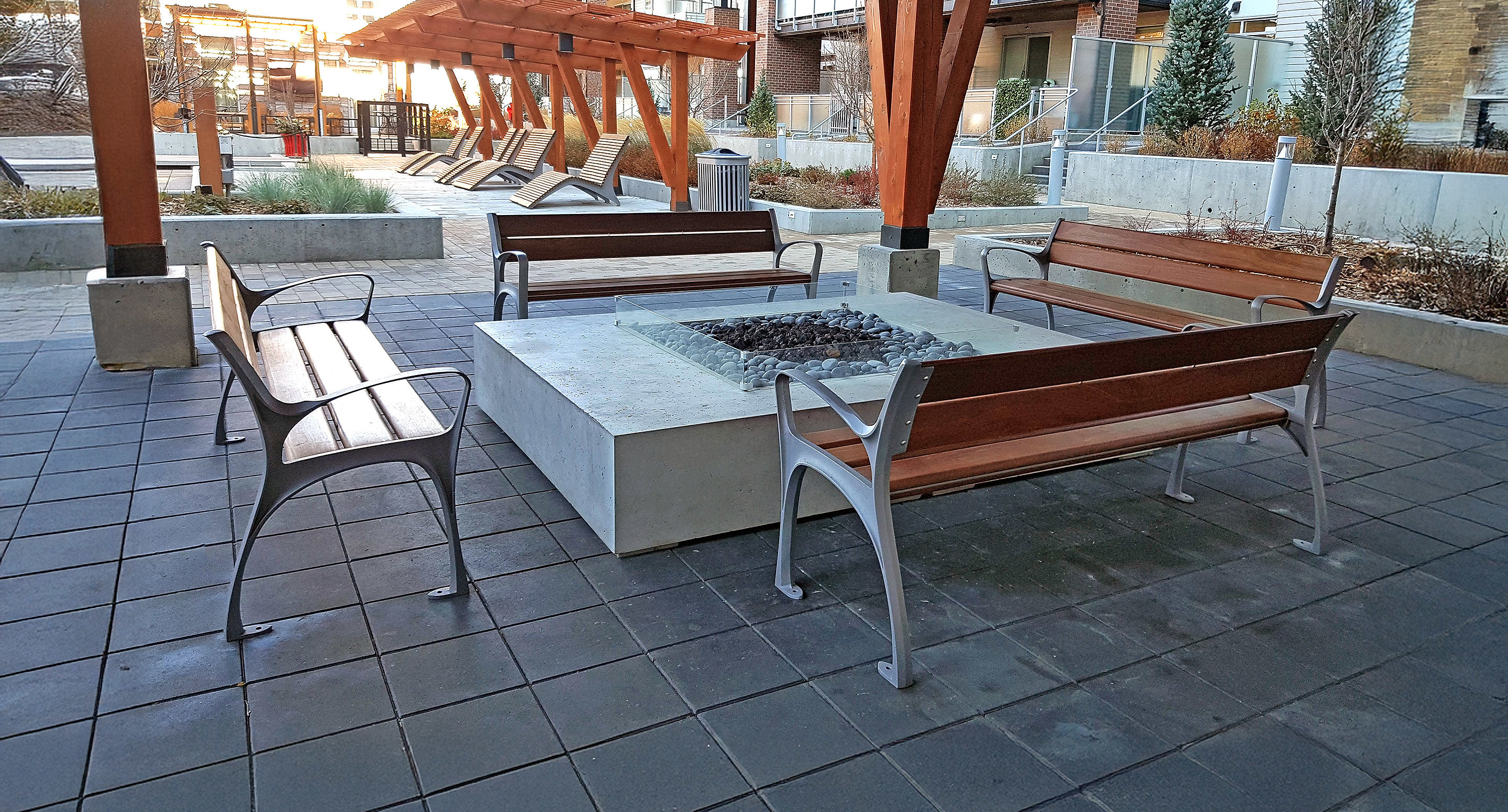 Four wooden benches surround a square concrete fire pit on a tiled patio, with a pergola and landscaped plants in the background.