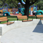 Three wooden benches are placed on a curved concrete wall near a paved area, with a colorful playground and green trees visible in the background.