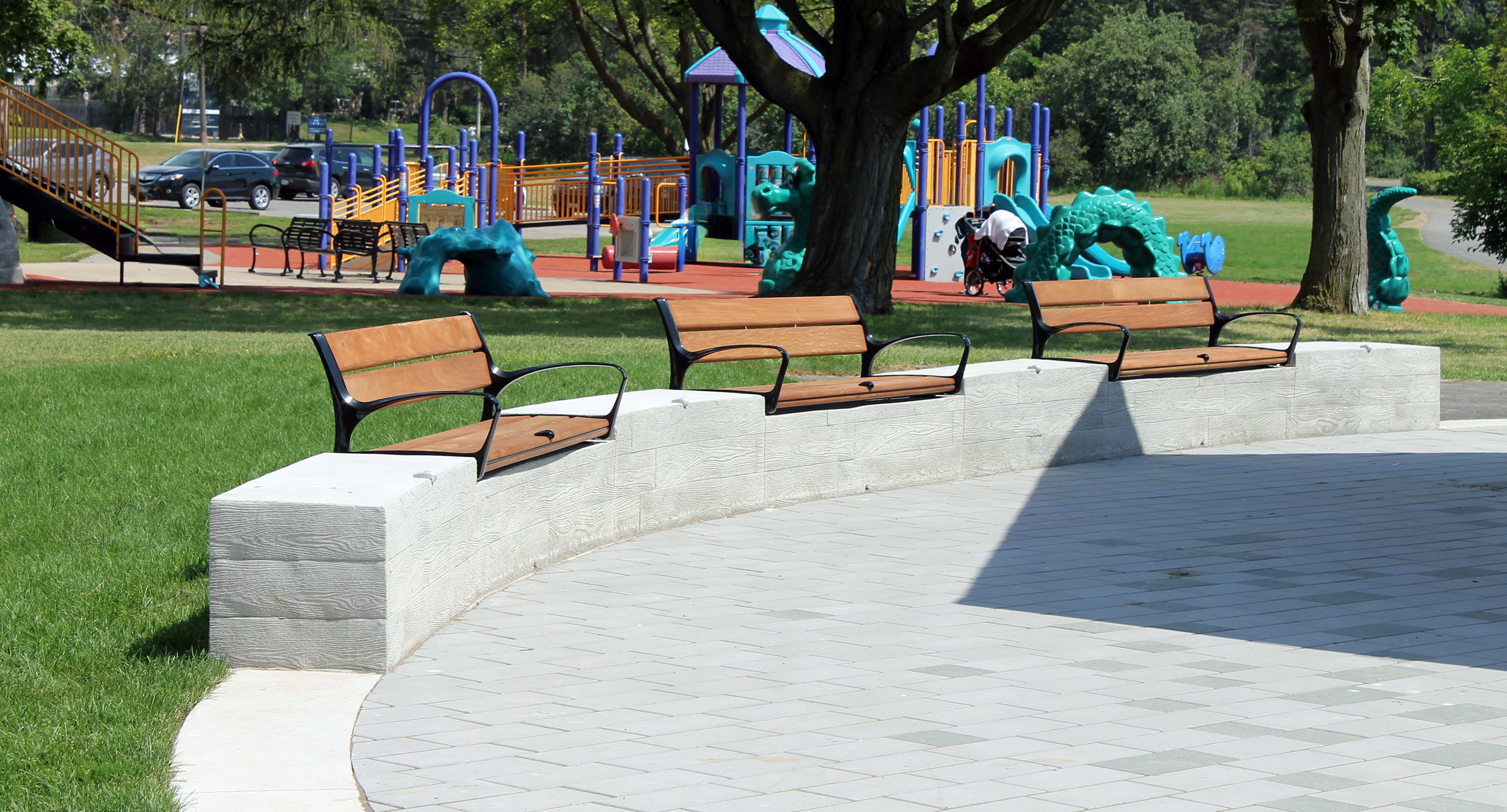 Three wooden benches are placed on a curved concrete wall near a paved area, with a colorful playground and green trees visible in the background.