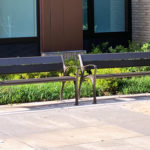 Two empty metal benches are placed on a paved area in front of a building with windows and landscaping.