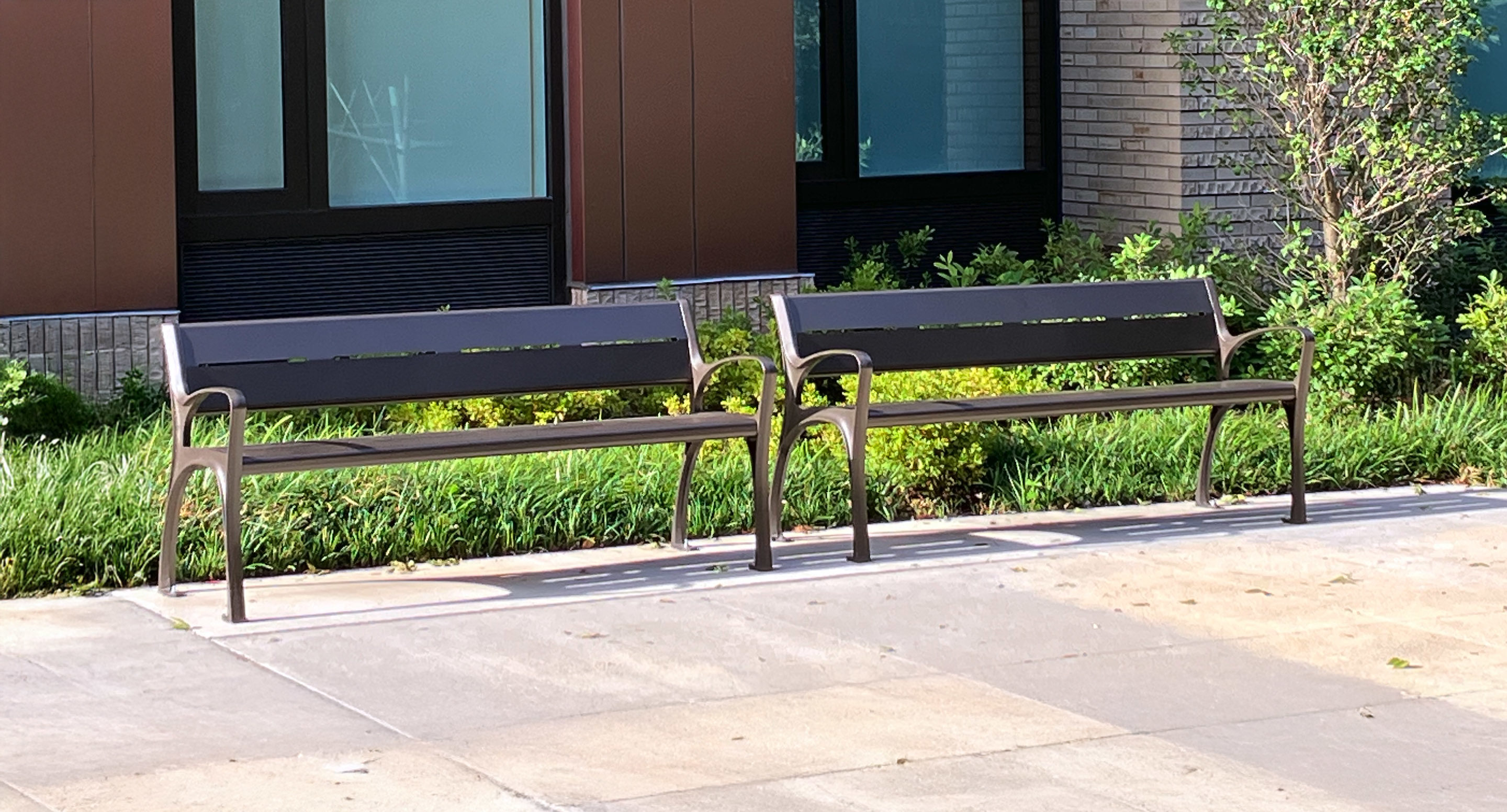 Two empty metal benches are placed on a paved area in front of a building with windows and landscaping.