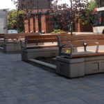 Three wooden benches with metal armrests are arranged along a paved urban sidewalk near a bus shelter, with a bus stopped in the background.
