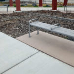 Gray metal bench with armrests sits on a concrete pad next to a mulched planting bed and a paved walkway, with red columns and a road in the background.