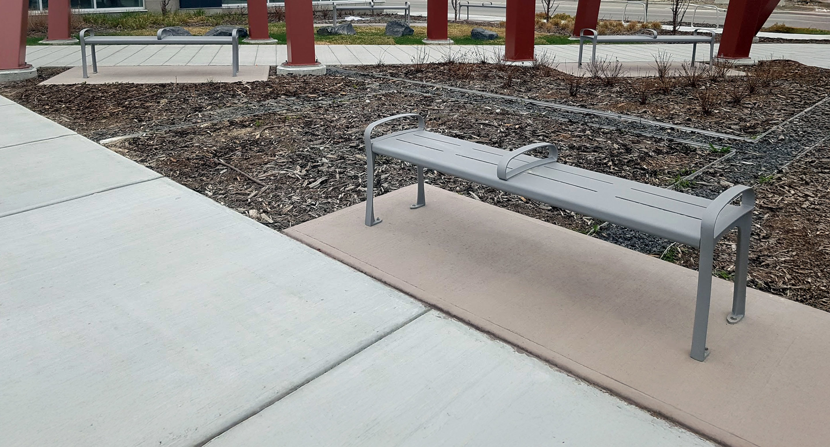 Gray metal bench with armrests sits on a concrete pad next to a mulched planting bed and a paved walkway, with red columns and a road in the background.