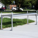 A modern metal bench with armrests stands on a concrete surface beside a patch of grass in a sunny outdoor park area.