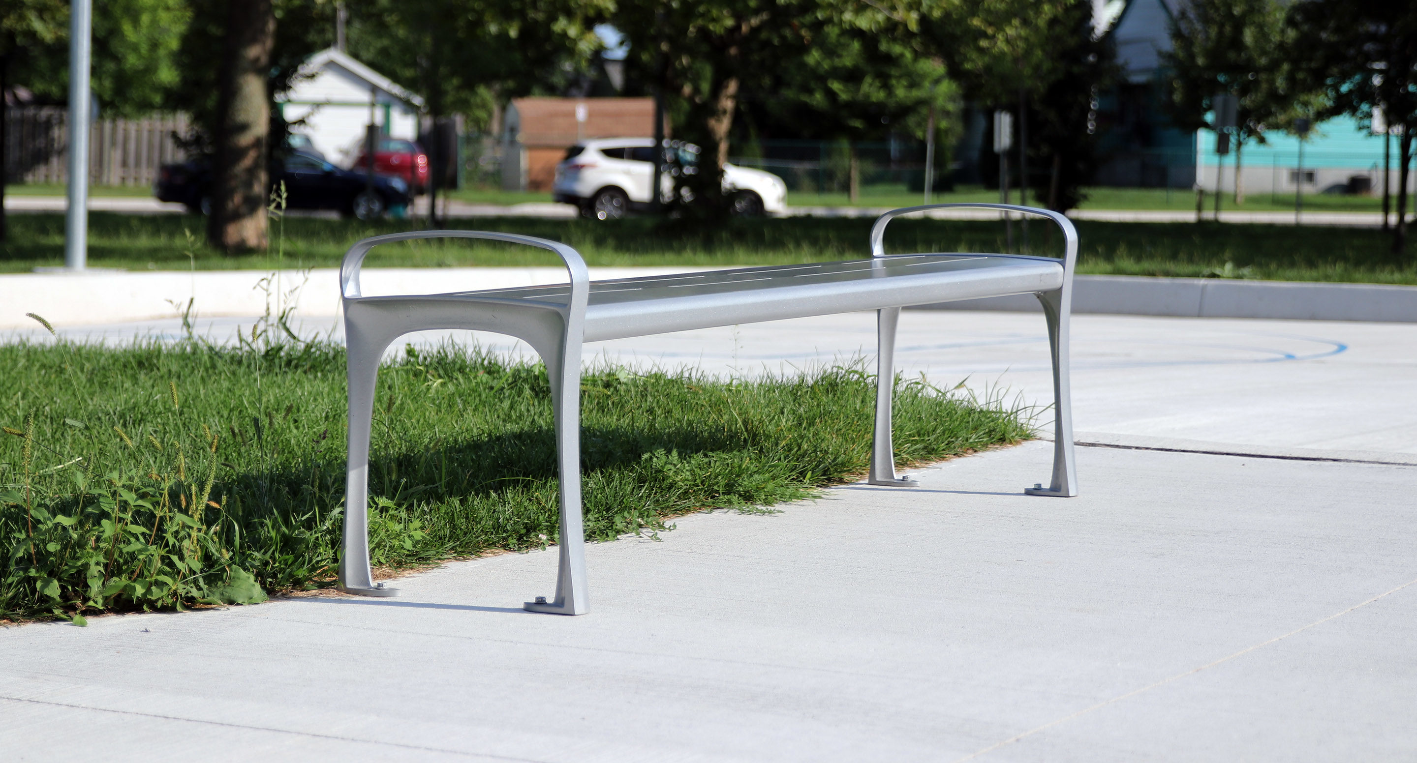 A modern metal bench with armrests stands on a concrete surface beside a patch of grass in a sunny outdoor park area.
